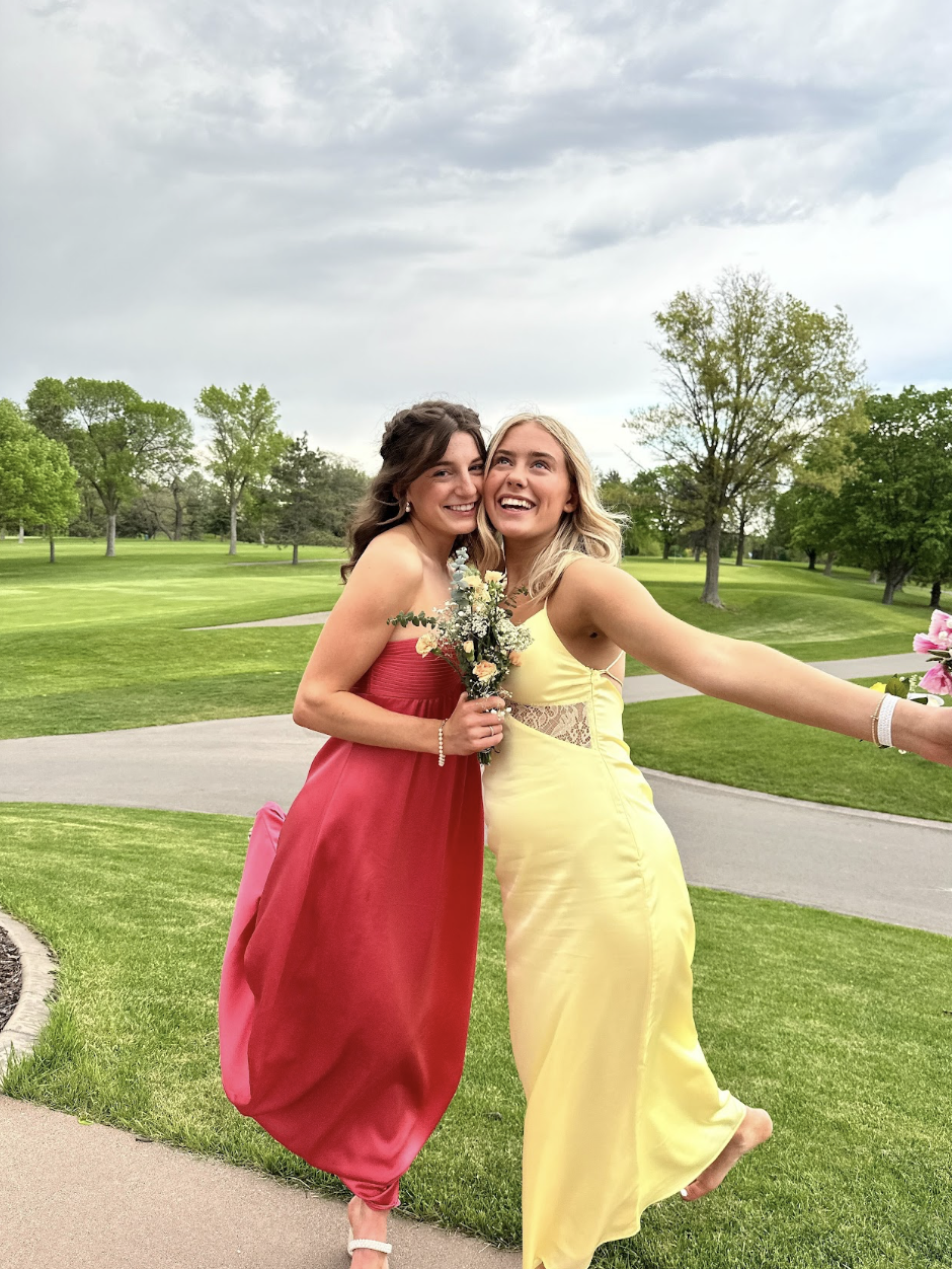 Two young women in formal dresses embracing outdoors on a grassy park with trees and cloudy sky.