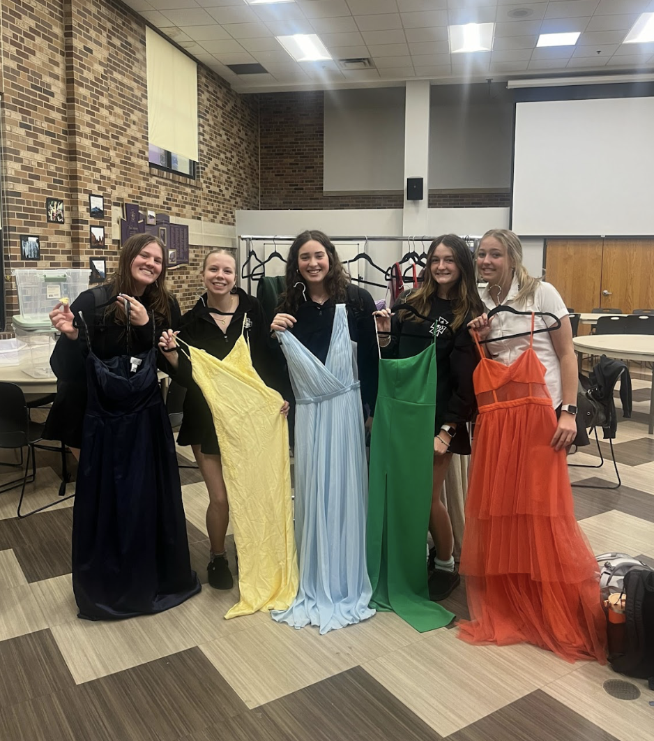 Five young women standing in a room, holding colorful dresses on hangers, smiling at the camera.