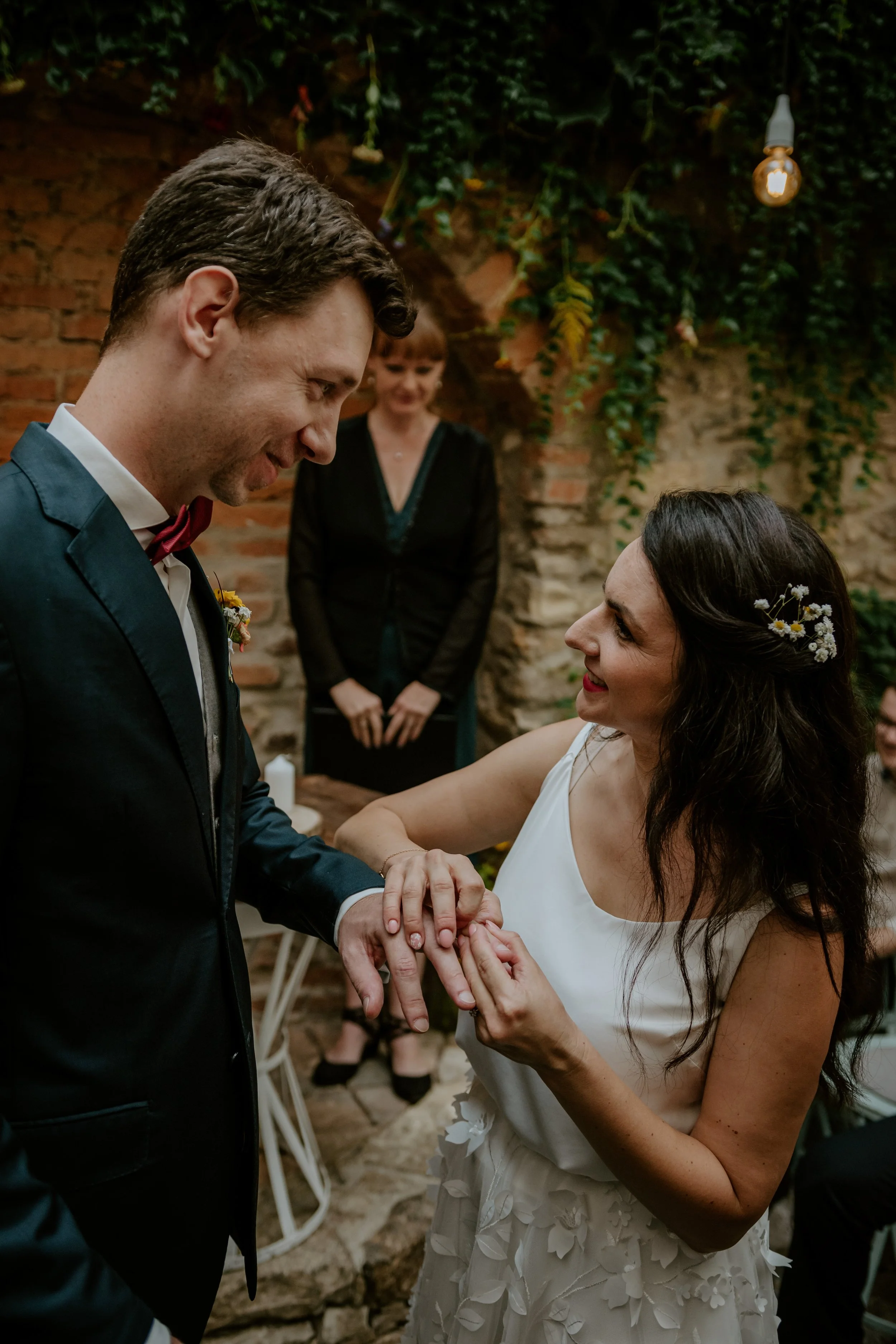 Žena v bílých šatech nasazuje prsten na mužský prst během svatebního obřadu, v pozadí sleduje host. Svatební fotografie, 