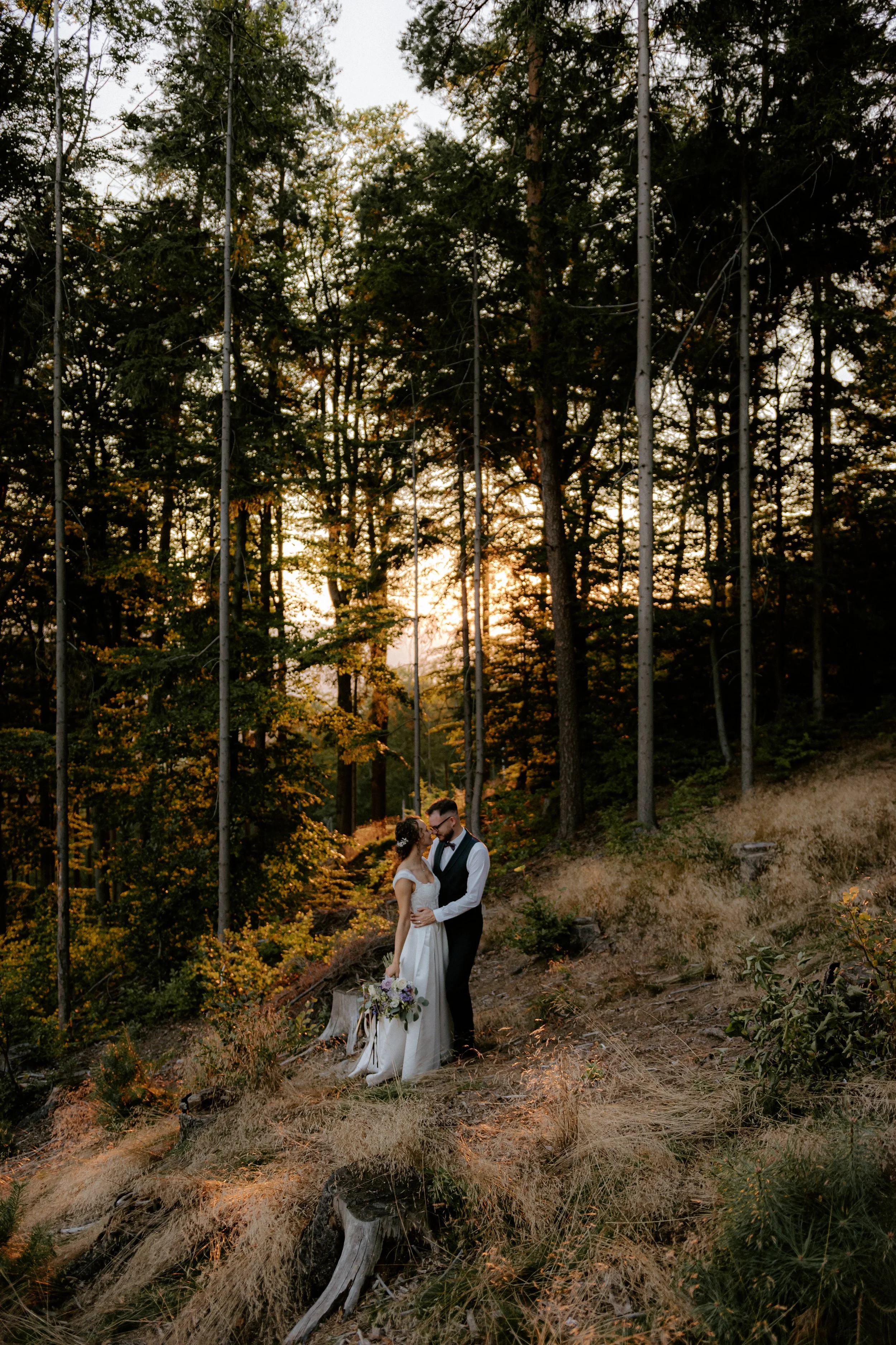 Nevěsta a ženich se objímají na zalesněném svahu při západu slunce, obklopeni vysokými stromy a suchou trávou, nevěsta drží kytici květin. Svatební fotografie, fotografové