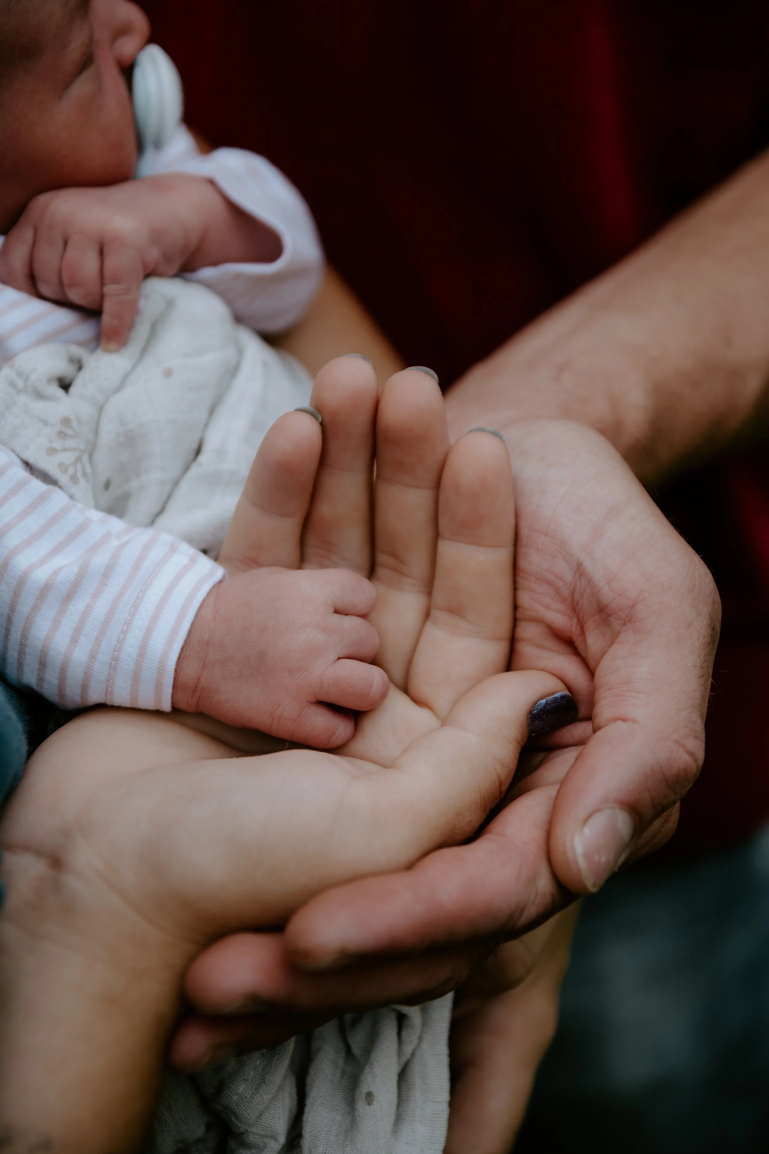 Detailní záběr novorozeného miminka s nalakovanými nehty, držícího ruku dospělé osoby, přičemž druhá dospělá ruka jemně podpírá ruku miminka. portrétní fotografie, novorozenecké focení, newborn
