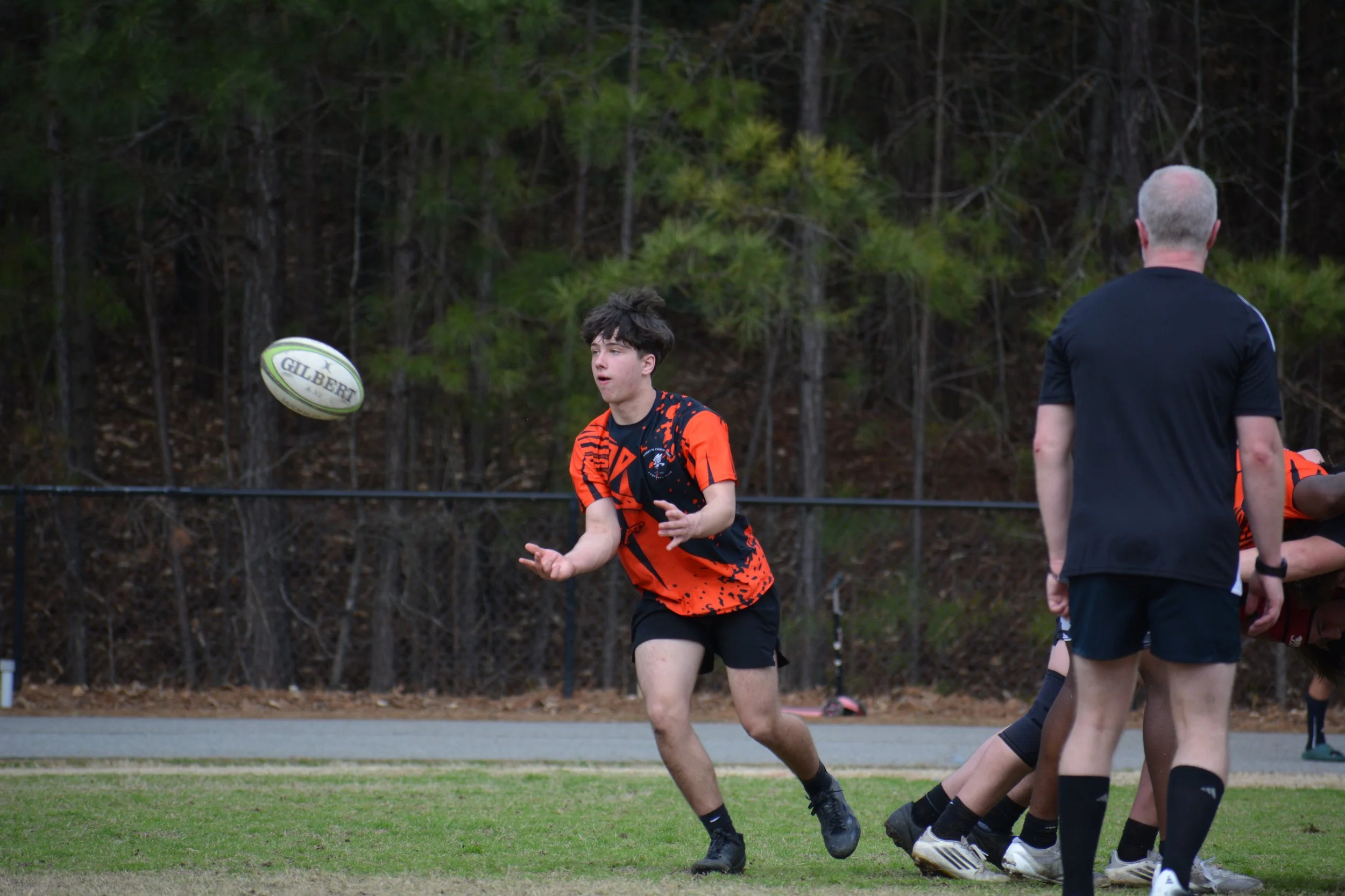 A high school boy playing rugby pitches the ball out of a scrum to his winger to run the ball down the field. The Sir or referee watches the play.