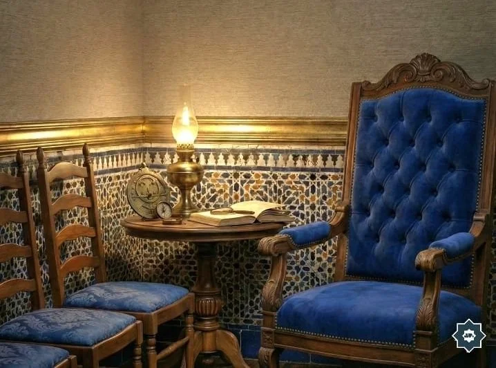 Hyperrealistic still life of a counsel-themed study at night featuring a large diamond-tufted navy silk armchair and three antique ladder-back chairs gathered around a lit brass oil lamp on a carved wood pedestal table. Nafsychology CE#3.