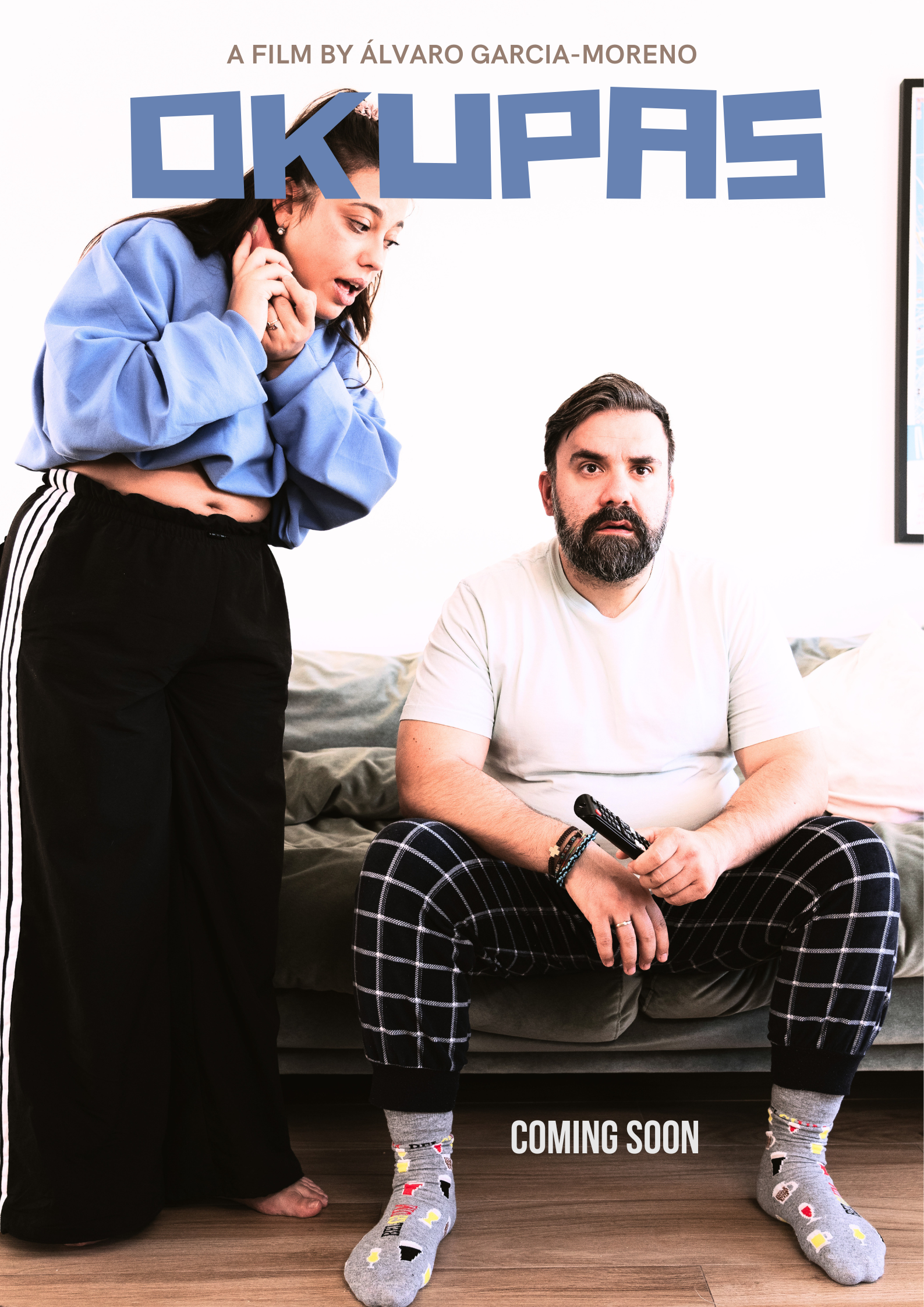 Film poster for “Okupas,” showing a woman in a blue sweatshirt standing beside a bearded man holding a TV remote, with text reading “A film by Álvaro Garcia-Moreno” and “Coming soon.”