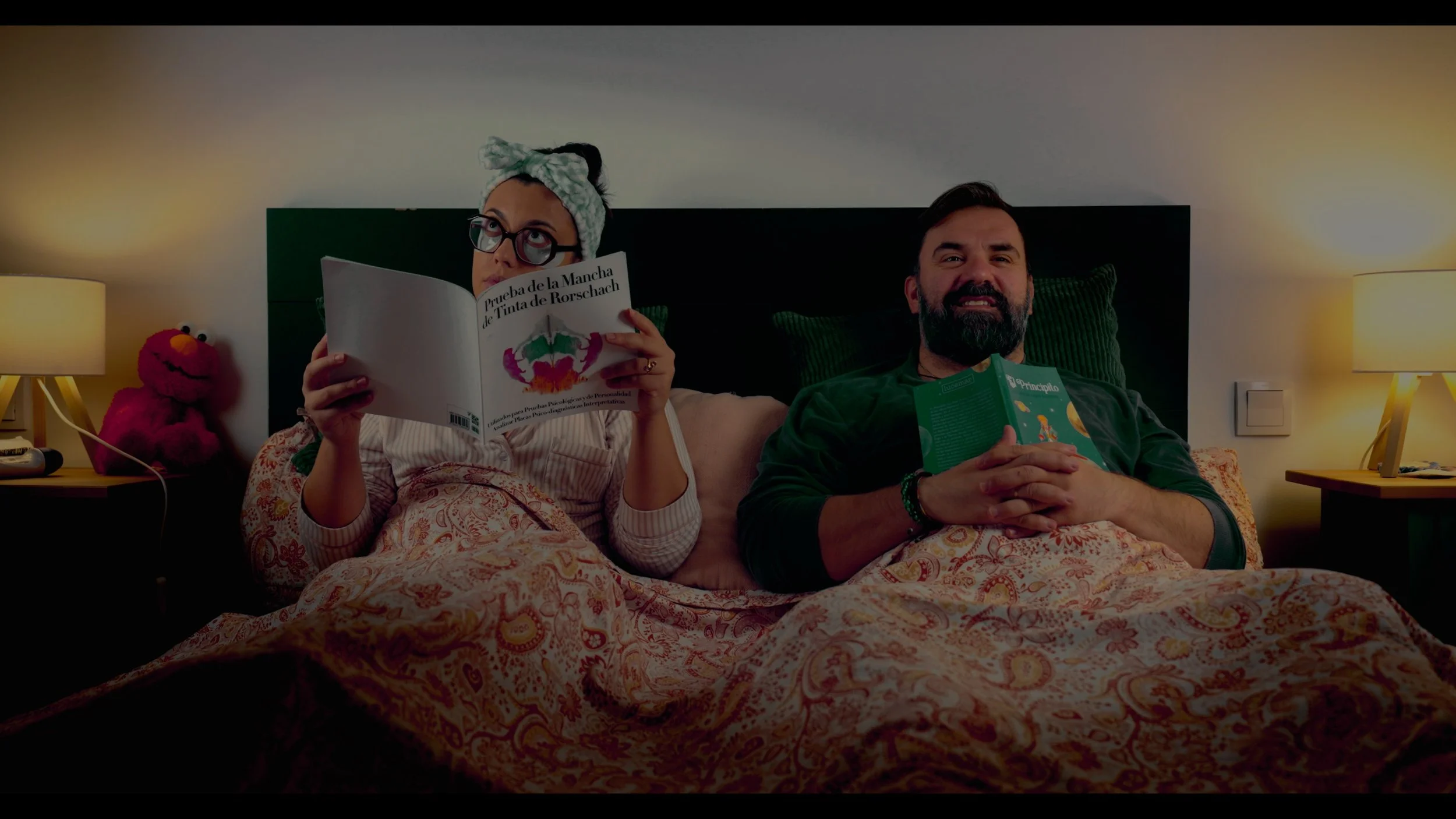 Couple reading books in bed under a patterned duvet, with bedside lamps and a red Elmo plush toy.