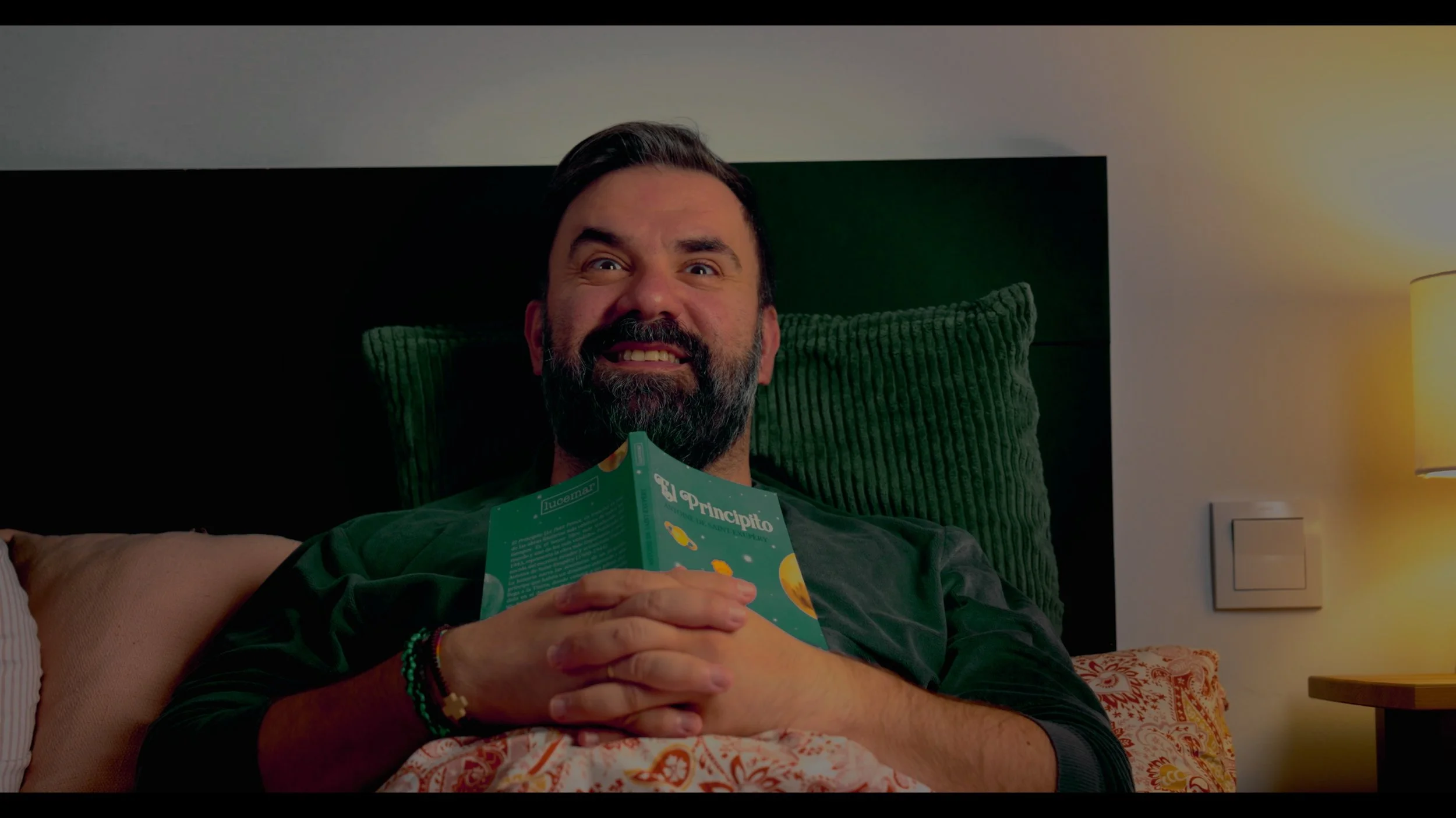 Bearded man sitting in bed and reading a green illustrated book under warm lamp light