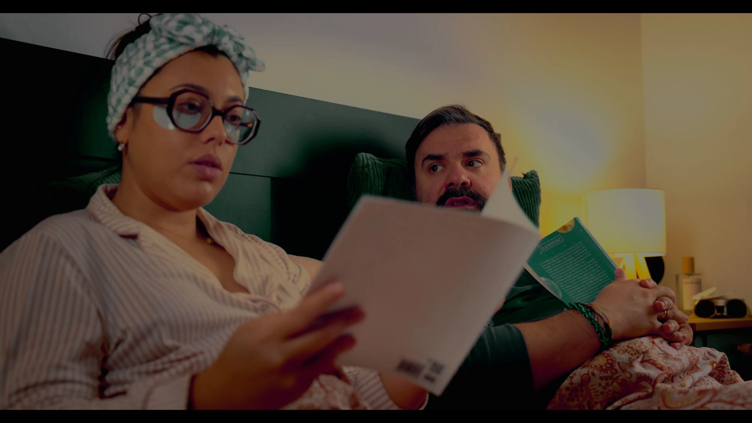 Woman wearing glasses and a headscarf reads papers while a man holds a green book in bed.