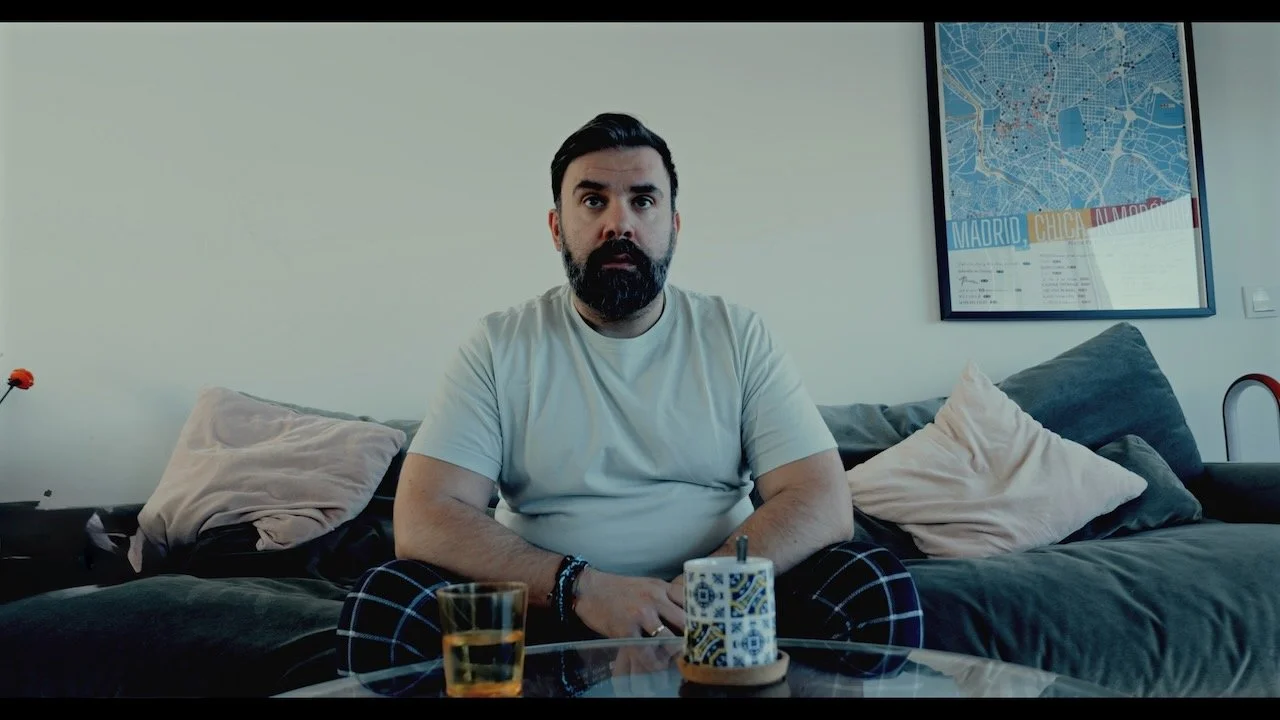 Bearded man in a white T-shirt sitting on a couch with a drink and patterned mug on a glass coffee table