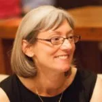 A woman with gray hair and glasses smiling, wearing a black top, sitting indoors with wooden walls in the background.