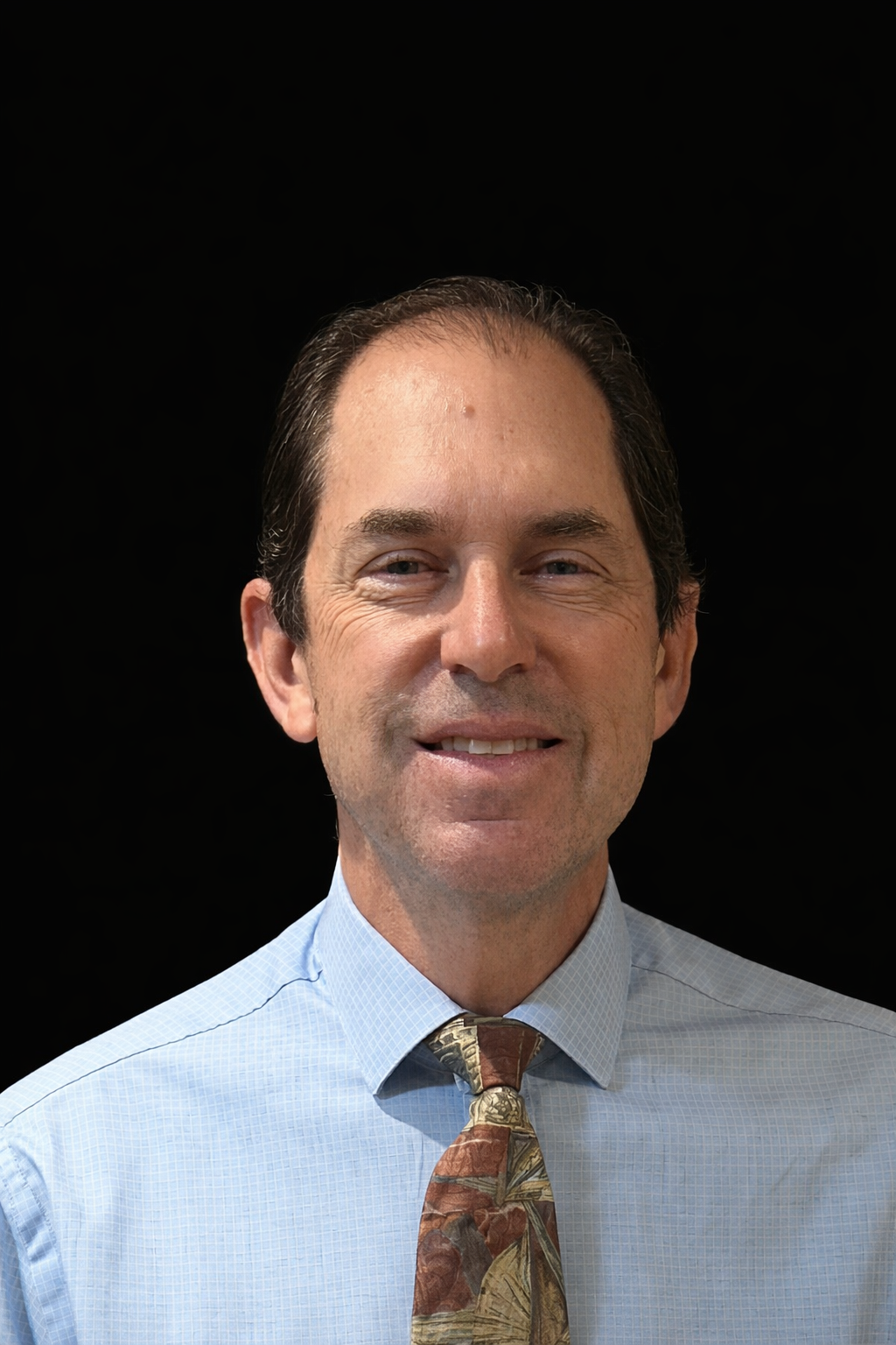 Portrait of a middle-aged man wearing a light blue collared shirt and a patterned tie, smiling, against a black background.