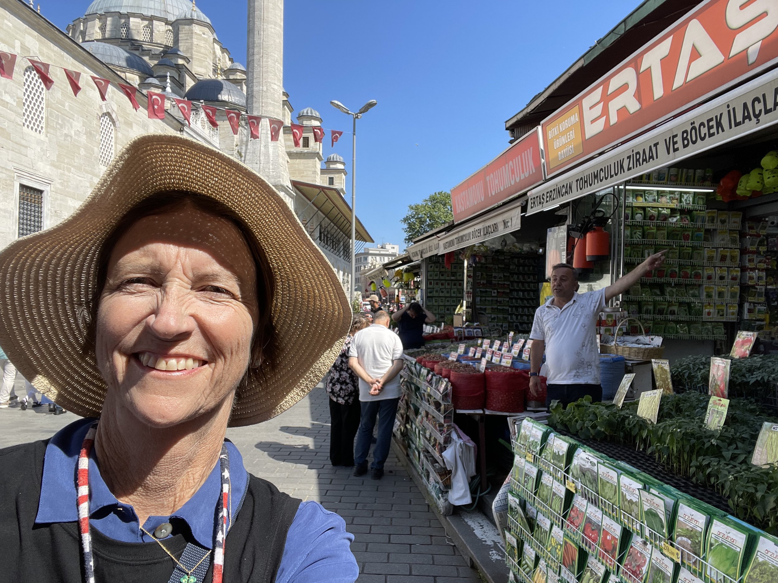 A woman wearing a large straw hat takes a selfie at an outdoor market in front of a traditional mosque with a tall minaret and domes, Turkish flags hanging, and market stalls selling various plants and gardening supplies.