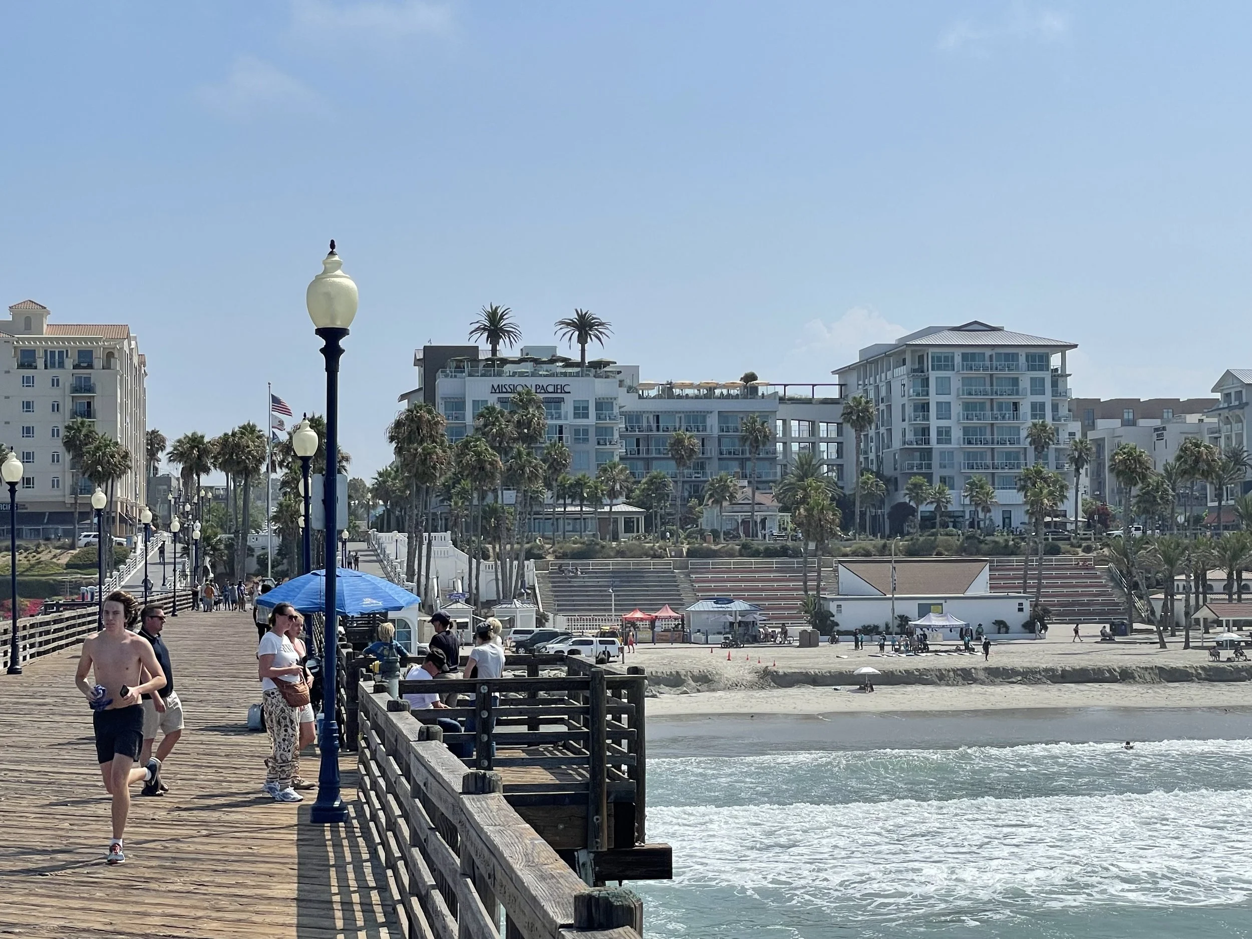A lively seaside boardwalk with people walking and relaxing, lined with vintage-style lamp posts, leading to a sandy beach with waves, and modern multi-story buildings with balconies and palm trees in the background under a bright blue sky.