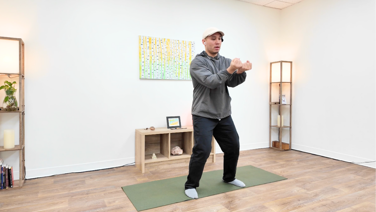 Man in gray hoodie and black pants doing a stretching exercise on a green yoga mat in a room with wooden flooring and minimalist decor.