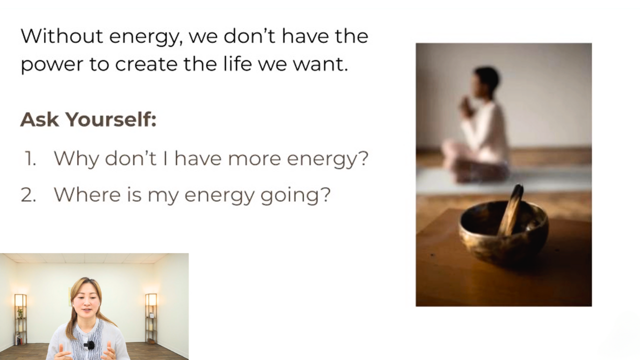 A woman practicing meditation or yoga in a peaceful room with minimal lighting, seated on the floor with hands in prayer pose, with a bowl and a wooden stick on a table in the foreground.