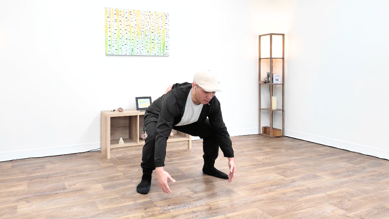 Person in black clothing and a white cap performing a yoga or stretching pose on a wooden floor in a room with minimal decor.