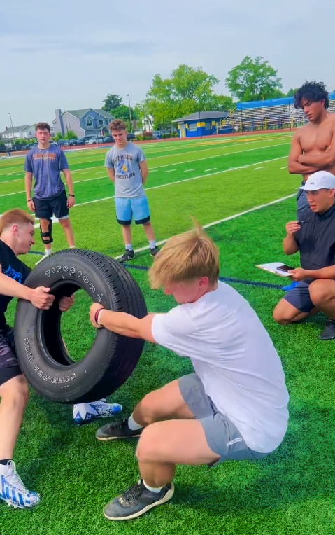 Two boys in athletic attire are participating in a tire-flipping activity on a sports field, with several other boys and a coach watching nearby.