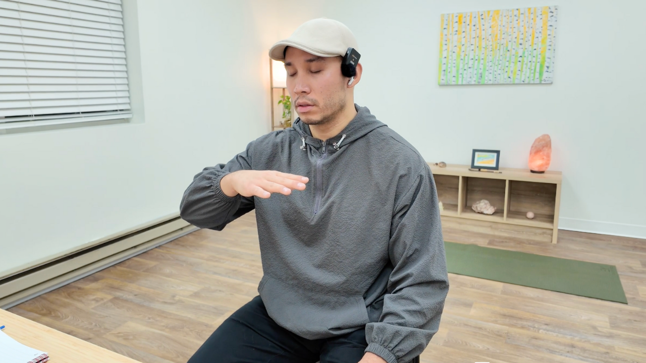 A man wearing a gray hoodie and beige cap, sitting with eyes closed. He has a headset with a microphone and is raising his right hand with the palm facing downward, in an indoor setting with a wooden shelf, abstract art, and a salt lamp in the background.