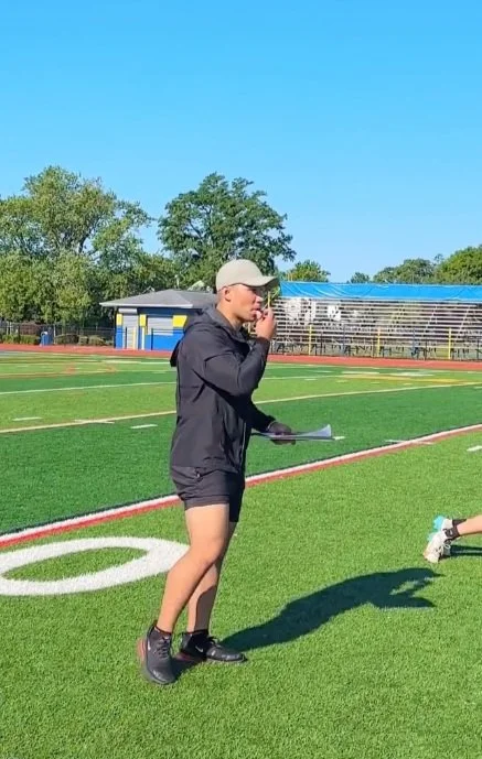 A woman on a football field, holding a clipboard and talking on a walkie-talkie, wearing a beige cap, black jacket, and shorts.