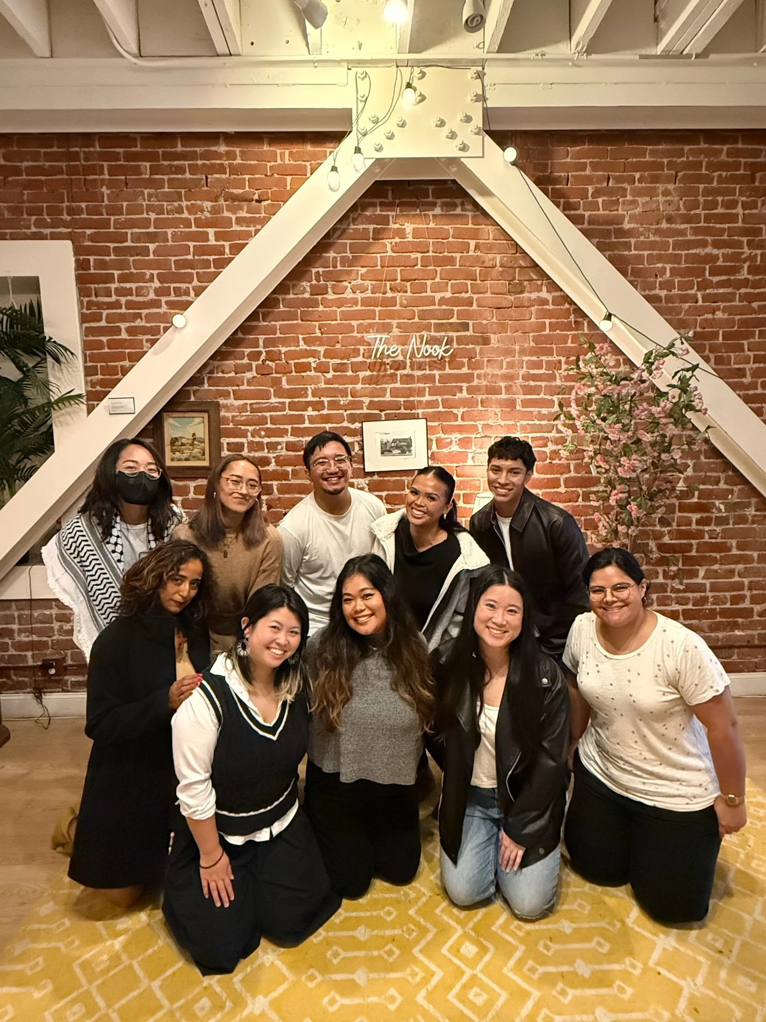 Group of ten diverse people posing for a photo indoors, in front of a brick wall with a sign that says 'The Nook,' decorated with framed photos and a flower arrangement.