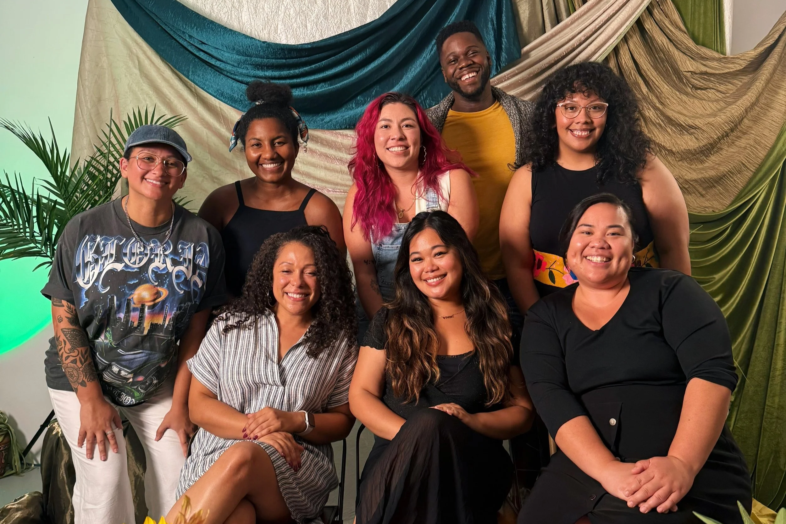 A diverse group of nine people, five women and four men, smiling and posing together indoors with colorful drapes and plants in the background.