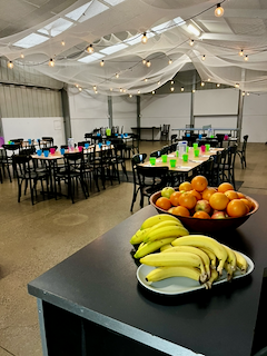 Banquet hall decorated with string lights and draped fabric ceiling, set up with tables and chairs, with a bowl of apples and bananas in the foreground.