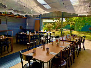Empty restaurant with tables and chairs, large windows showing trees and outdoor scenery, ceiling fans, and a blue wall.