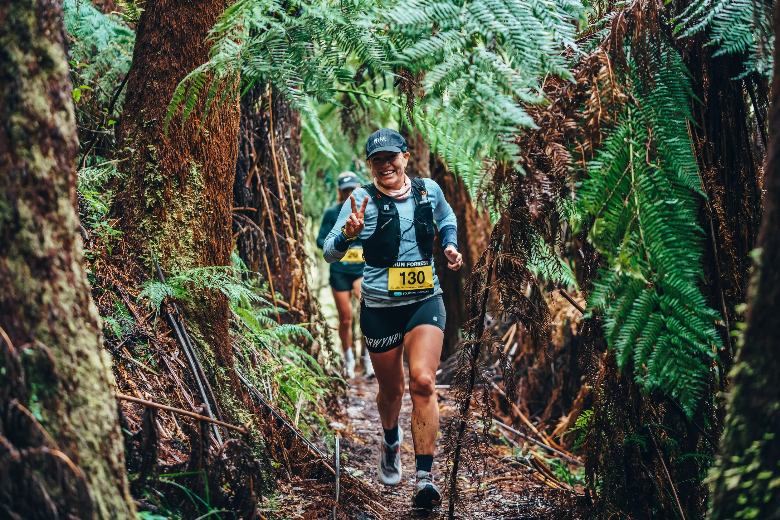 A woman running through a lush, green forest trail during a race, smiling and making a peace sign with her fingers, wearing a race bib number 130.