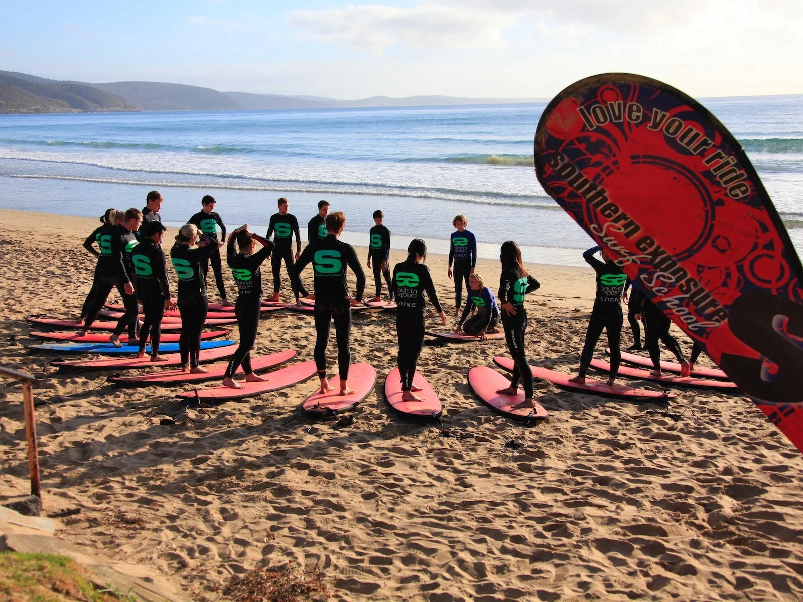 Group of people in wetsuits preparing for a surfing lesson In Torquay on a sandy beach, with surfboards laid on the sand and a large informational flag in the foreground.