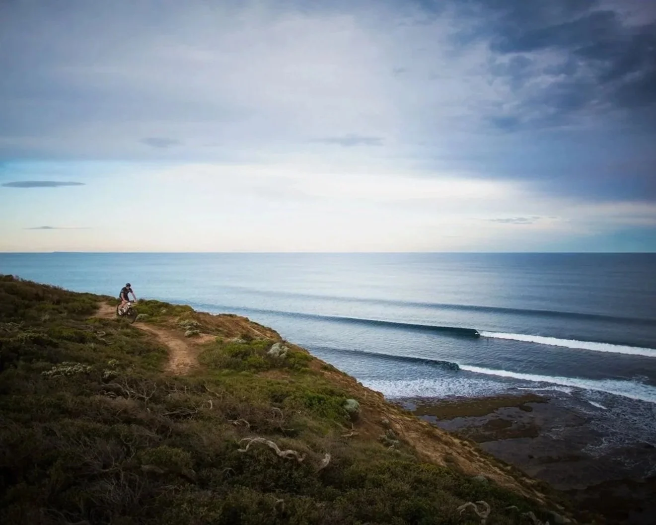 A person riding a bicycle on a dirt trail along a coastal cliffs above Bells Beach in Torquay with the ocean and cloudy sky in the background.