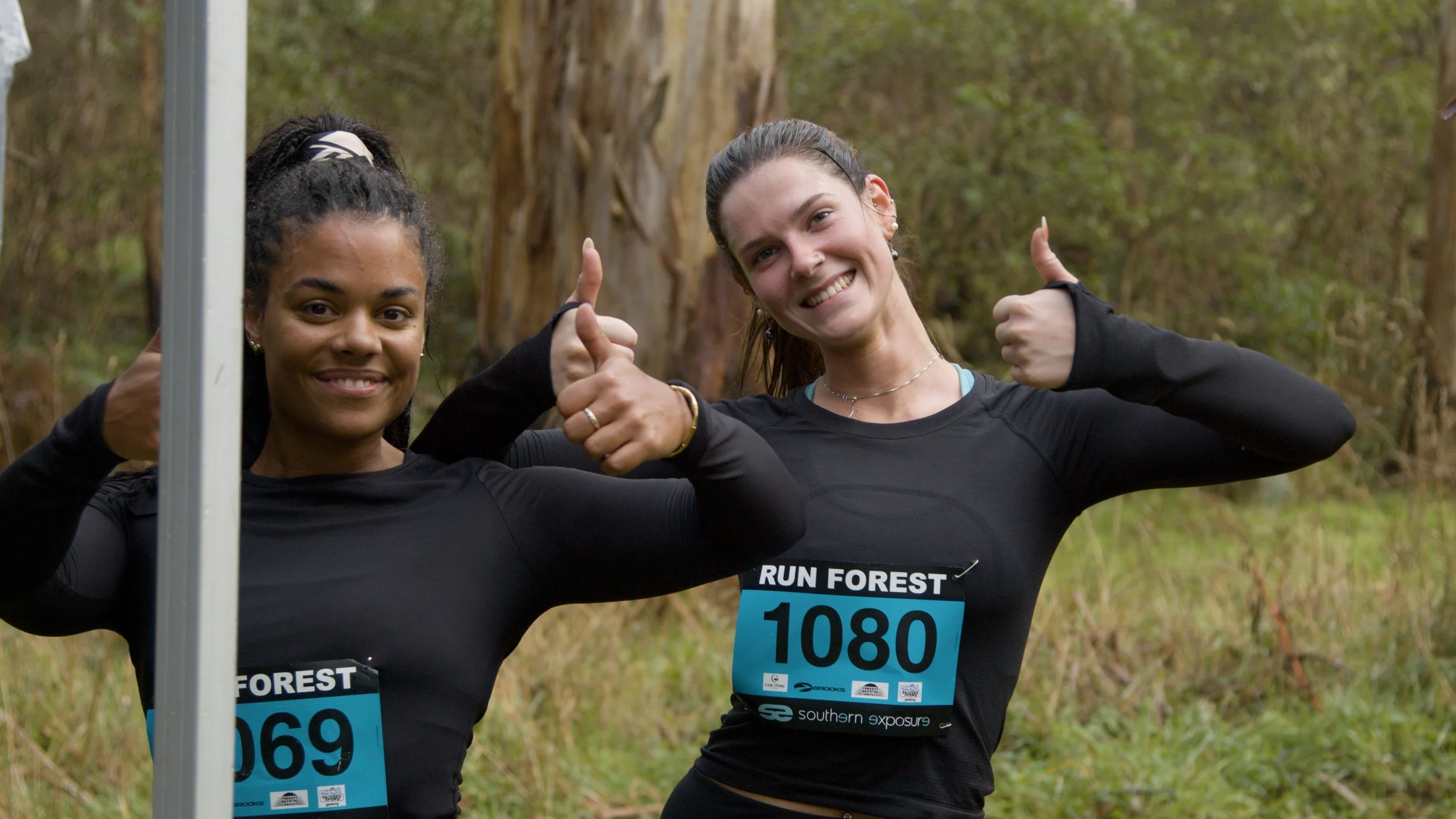 Two women smiling and giving thumbs-up gestures outdoors in a wooded area, wearing black athletic shirts with race bibs that say "RUN FOREST" and numbers 1089 and 1080.