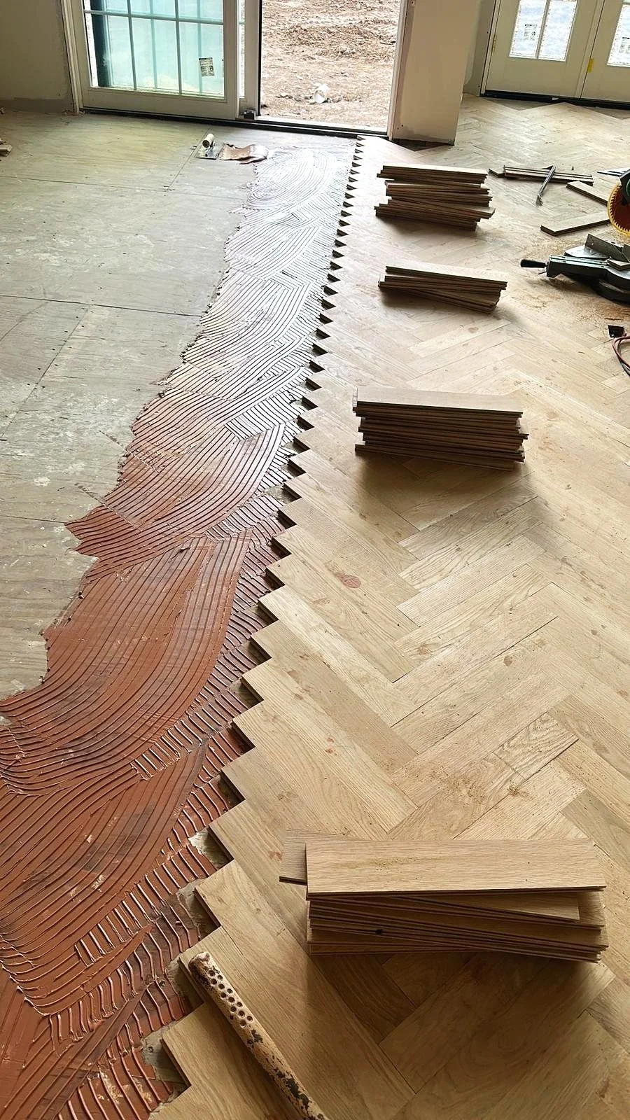 Interior of a room under construction, with a partially installed herringbone wood flooring. Piles of wooden planks are arranged along the edge, and tools are visible on the floor, with a sliding glass door leading outside.