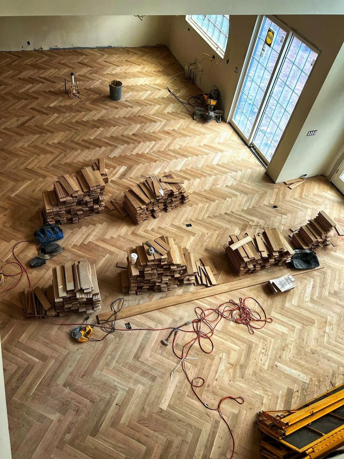 Construction site with newly installed hardwood flooring, wooden planks stacked in several piles, tools, and construction materials visible in the room.