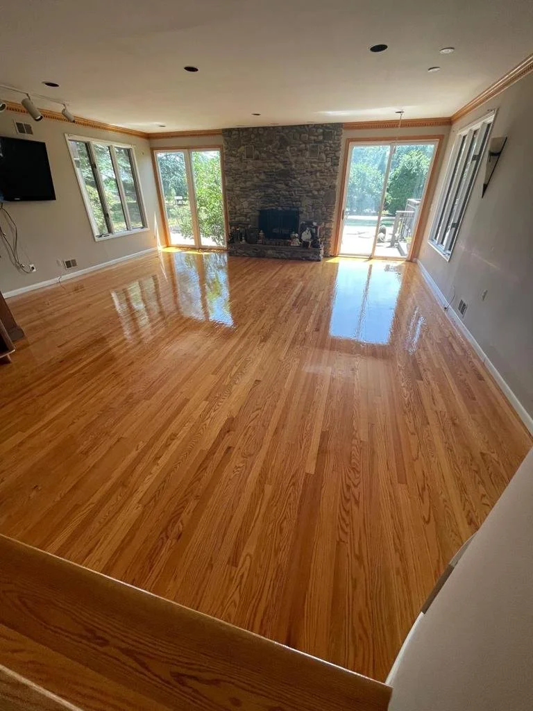 Empty living room with polished hardwood floors, a stone fireplace, large windows and sliding glass doors, and a mounted TV on the wall.