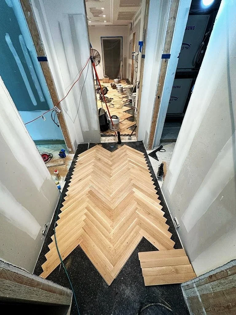 A hallway under renovation with new wood herringbone flooring being installed, construction tools and supplies scattered around, and walls partially finished.