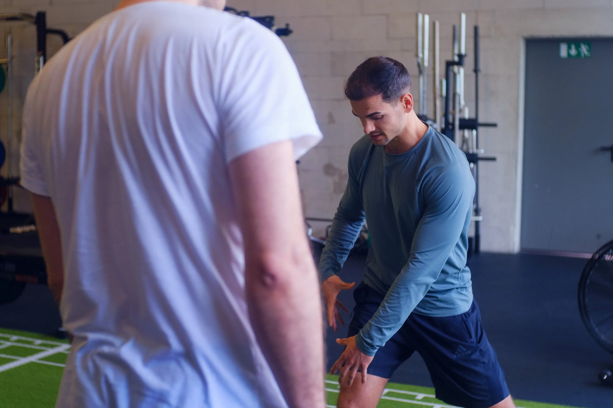 Ein Mann macht Dehnübungen in einem Fitnessstudio, während eine andere Person ihm zuschaut.
