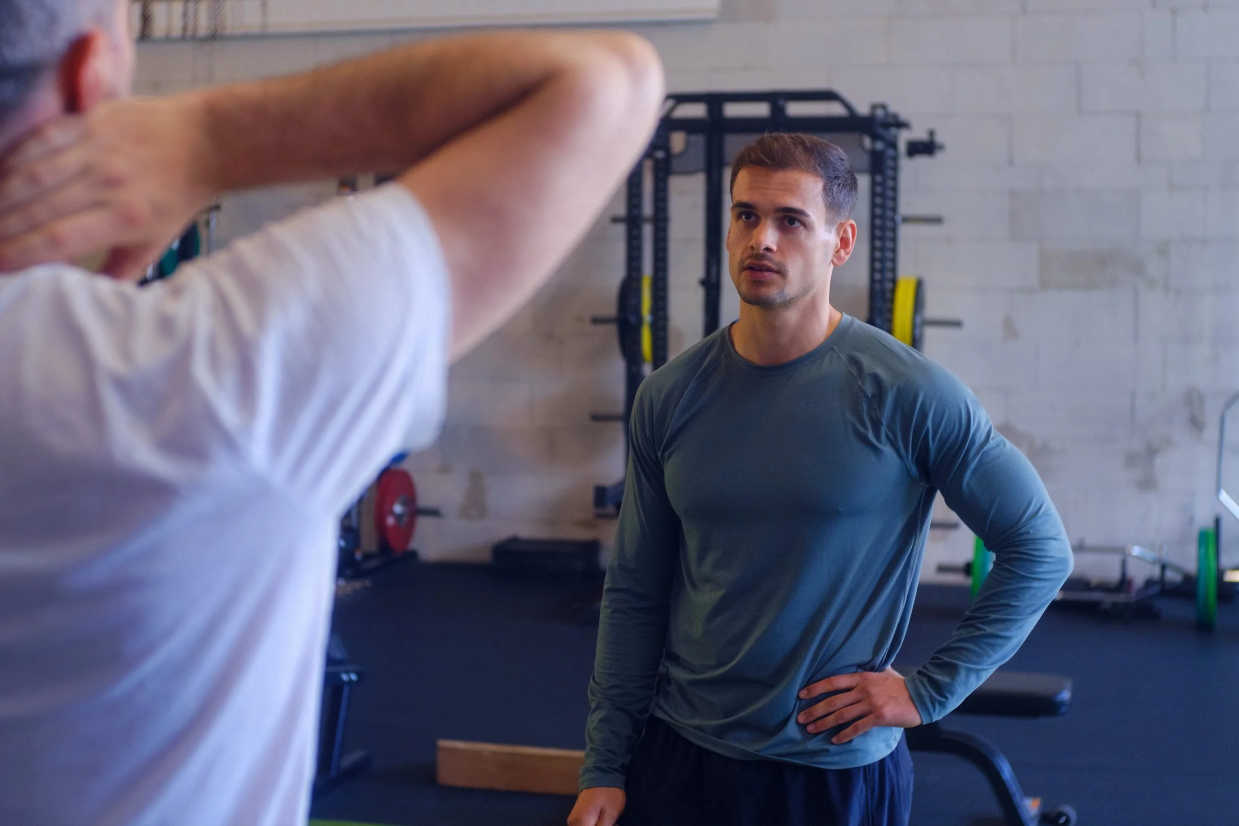 Ein junger Mann in einem Fitnessstudio hört auf einen älteren Mann, der ihn berät, mit modernen Fitnessgeräten im Hintergrund.