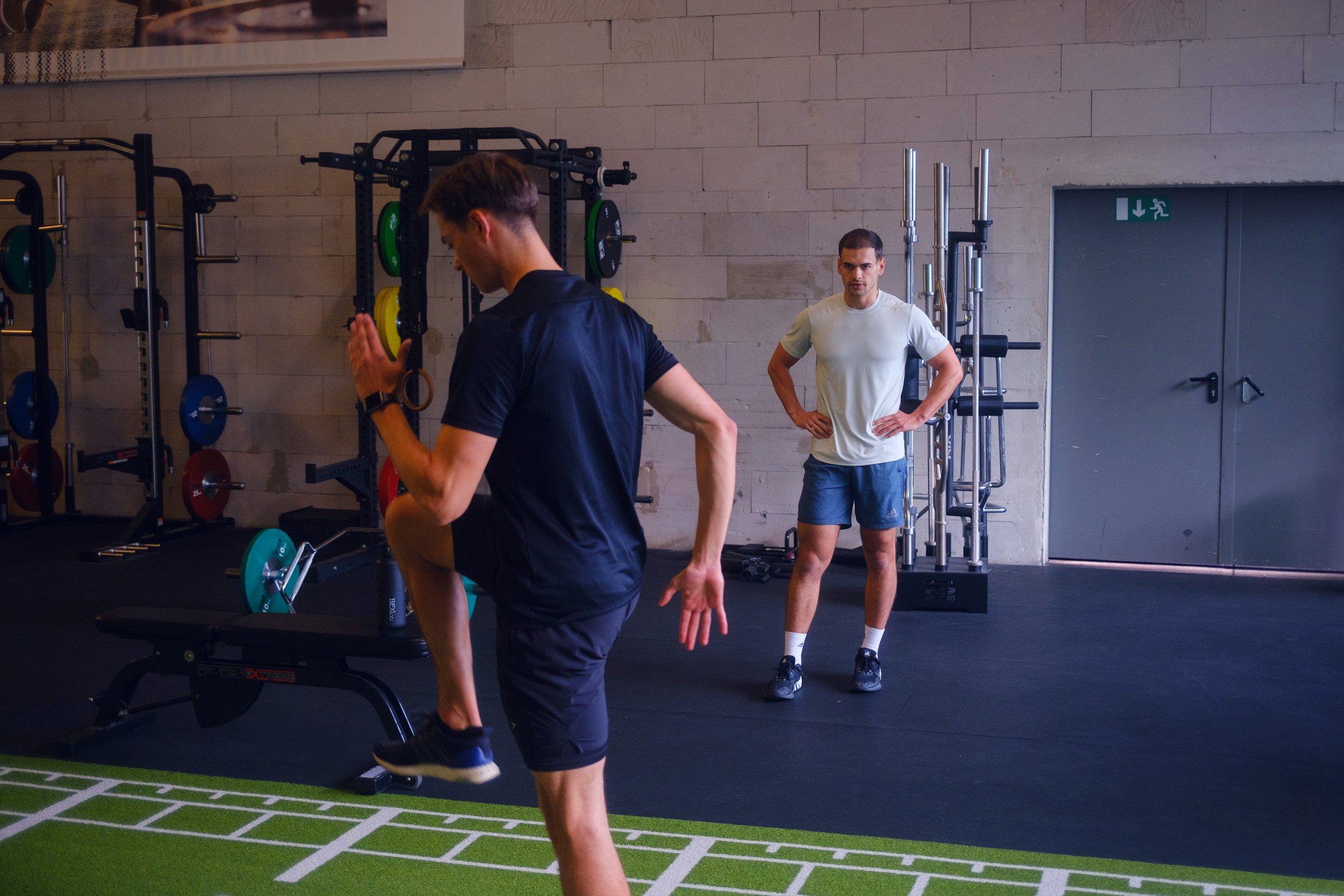Zwei Männer trainieren im Fitnessstudio. Der eine führt Kniebeugen auf einem Bein und der andere beobachtet. Im Hintergrund sind Gewichte und Fitnessgeräte sichtbar.
