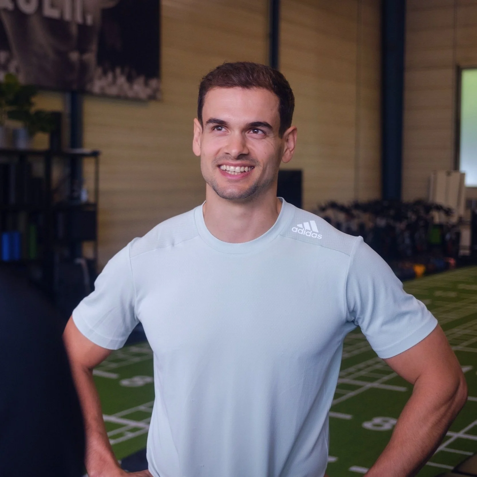 Ein junger Mann trägt ein weißes adidas-T-Shirt, steht in einem Fitnessstudio und lächelt.