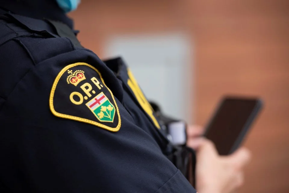 Close-up of a police officer's shoulder patch with the badge of O.P.P. (Ontario Provincial Police), showing a shield with a crown, a red cross, and a green background, while the officer looks at a smartphone device.