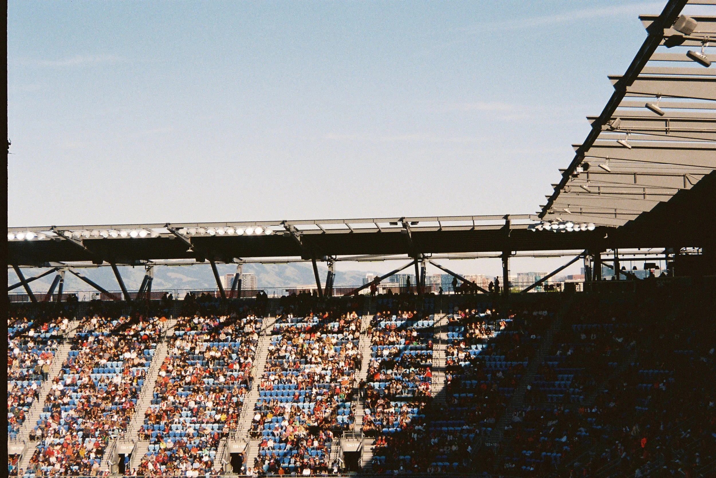 Crowd of spectators seated in a stadium with a partially covered roof, during daytime.