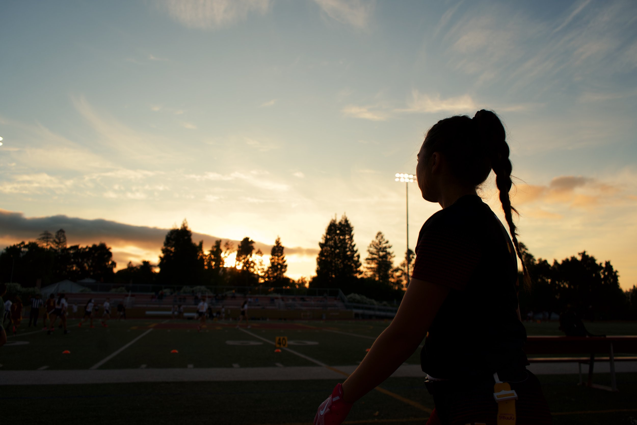 Silhouette of a girl in a sports uniform standing on a football field at sunset, with other players practicing in the background.