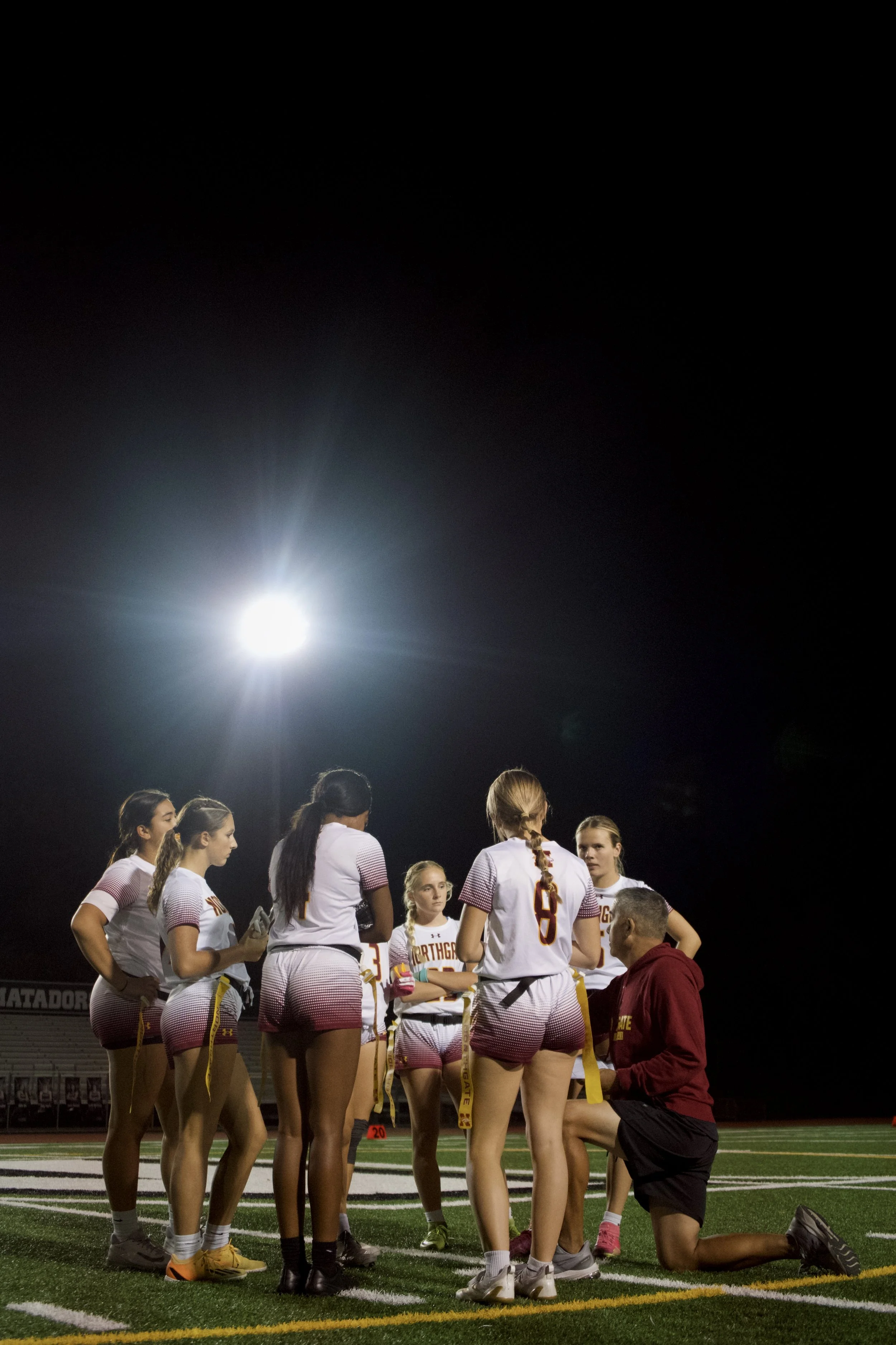 Girls' field hockey team gathered around coach on nighttime field under bright stadium lights.