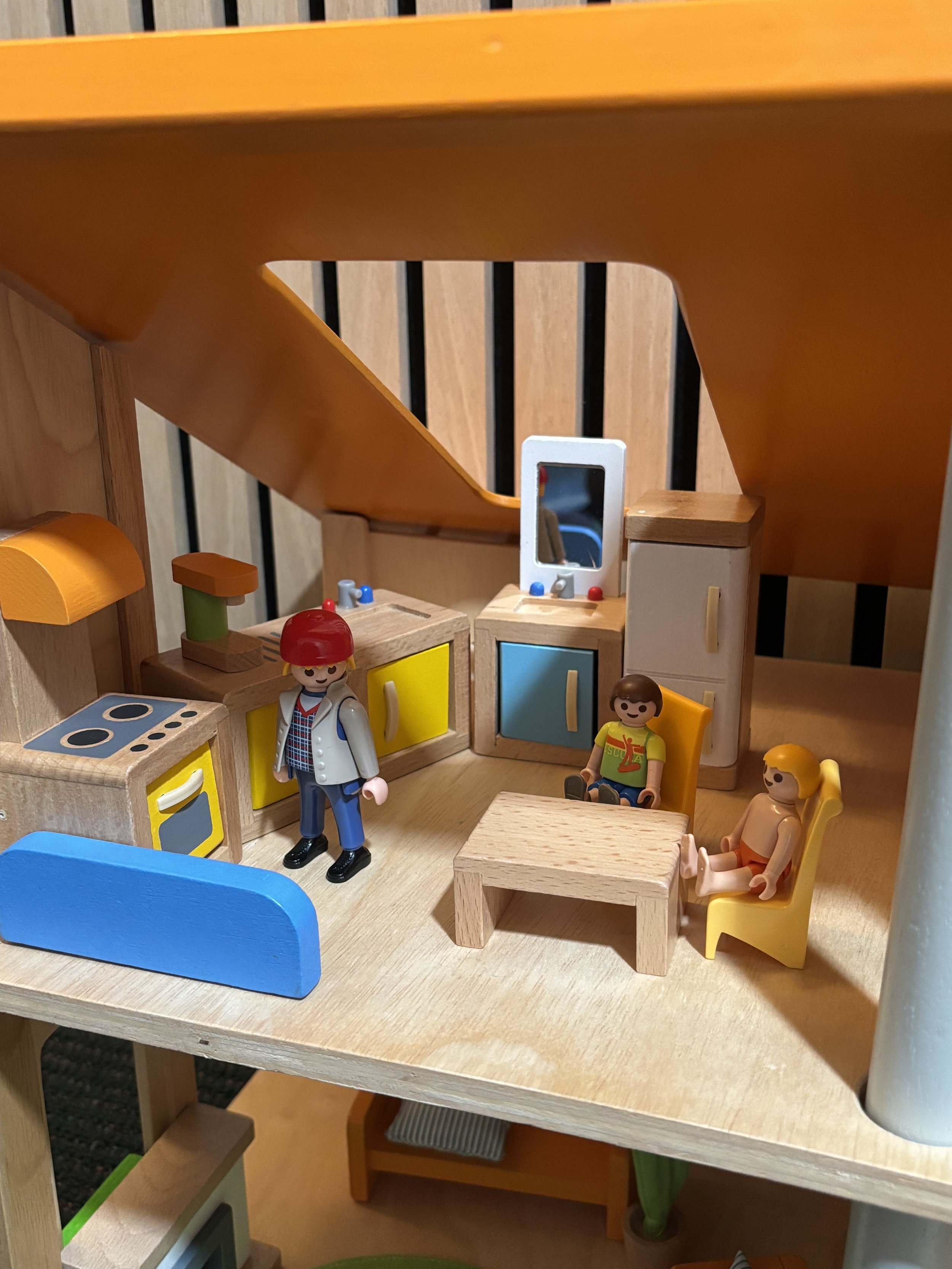 A wooden toy kitchen set with a yellow oven, a stove top, and a small mirror. There are plastic figurines of children, one with red hair and another with brown hair, sitting and standing near the kitchen.