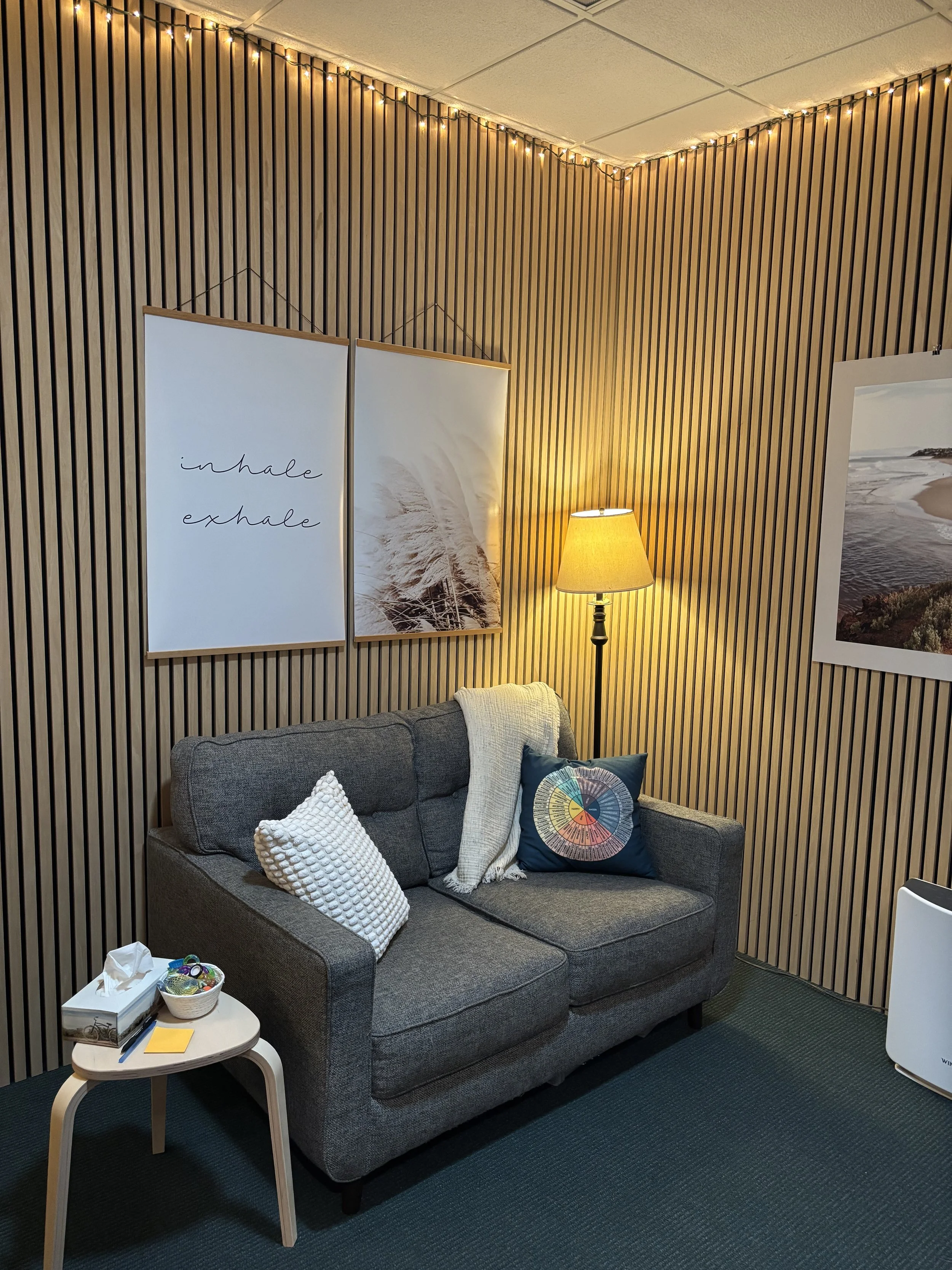 A cozy seating area with a gray sofa, decorated with white and patterned pillows, in a room with wood slat wall paneling. There is a small round side table with a bowl of miscellaneous items, a tissue box, and a yellow sticky note. A black floor lamp with a beige lampshade provides warm lighting, with additional string lights along the ceiling. Two framed pictures hang on the wall, one with the words ‘inhale exhale’ and a feather image, the other showing a beach scene.
