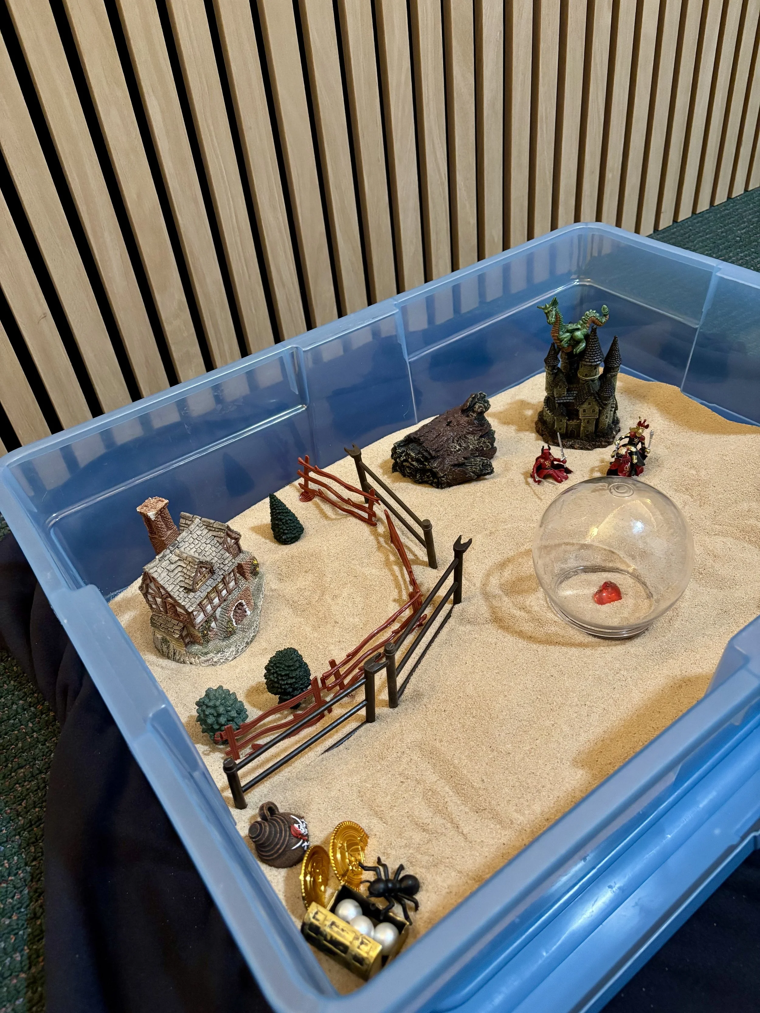 A therapy sand tray setup with miniature castles, trees, soldiers, and accessories in a blue plastic container.