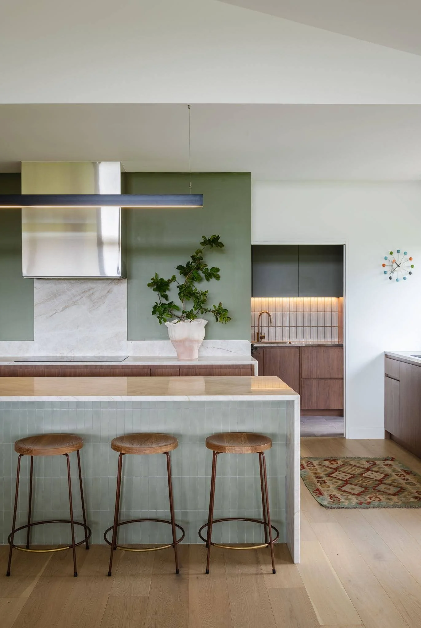 Modern kitchen with light green tiled island, three wooden bar stools, a green accent wall, a white pot with a leafy green plant, a built-in microwave and oven, a black and copper faucet, a patterned rug, and a decorative analog clock on the wall.