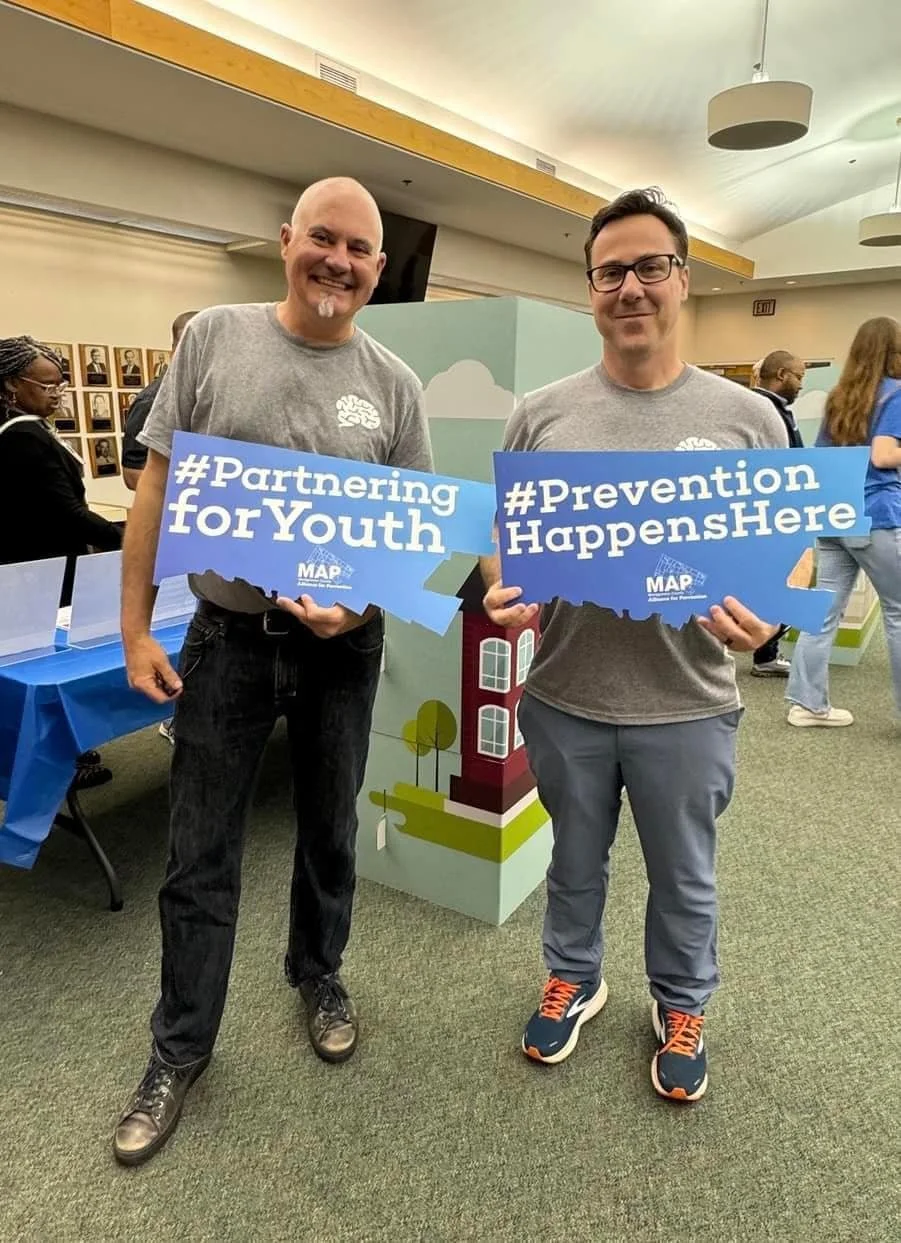 Two men wearing gray t-shirts standing indoors, holding signs that read '#Partnering for Youth' and '#Prevention Happens Here'.