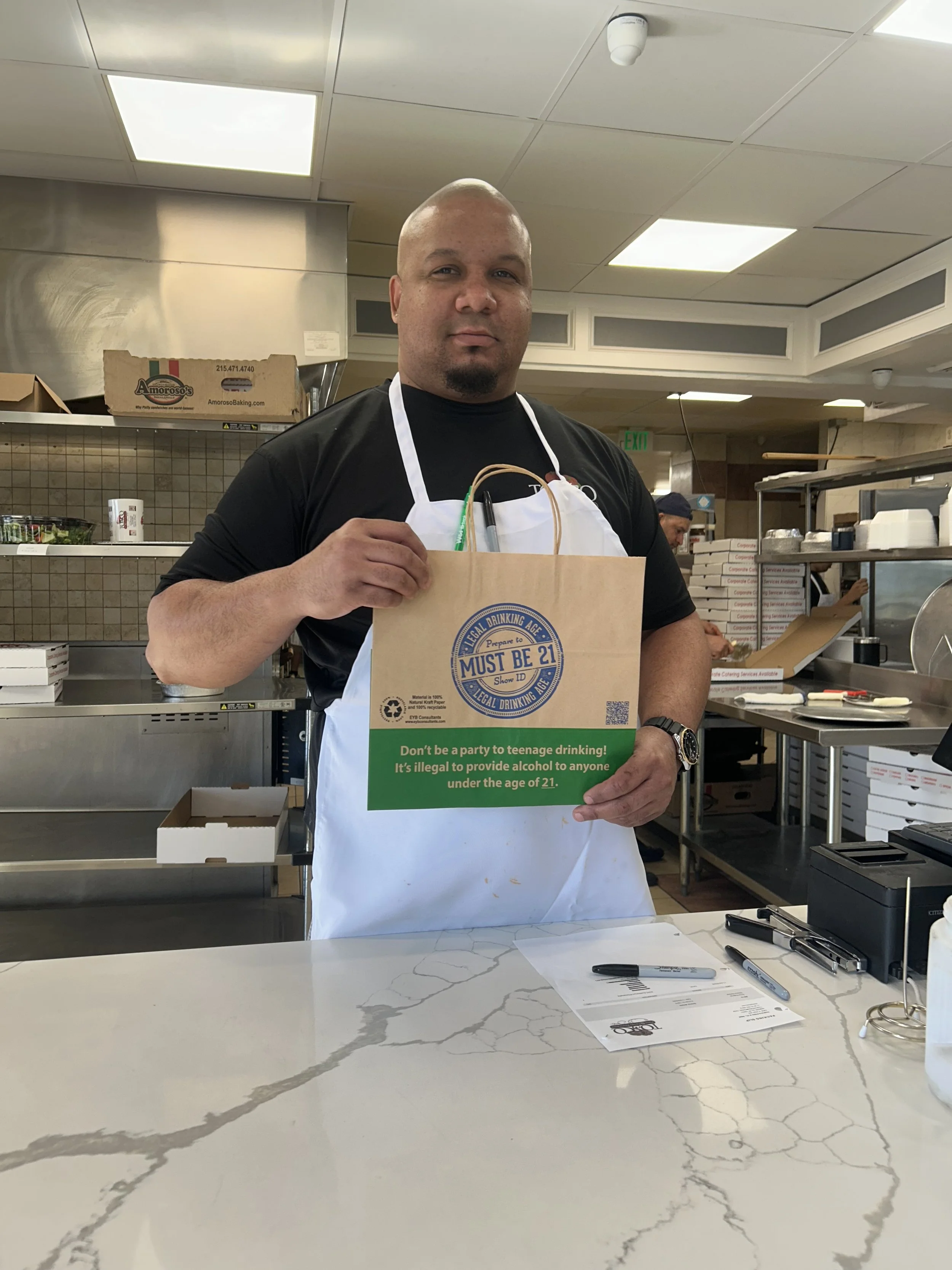 Man in a black shirt and white apron standing behind a white marble counter in a kitchen, holding a brown paper bag with a green label that reads, 'Don't be a party to teenage drinking! It's illegal to provide alcohol to anyone under the age of 21.'