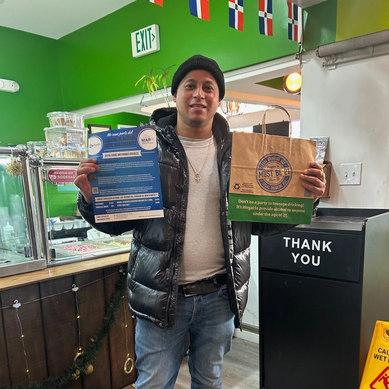 A man standing inside a restaurant or store, holding a blue social responsibility flyer in one hand and a brown paper bag with a green and black label in the other hand. The background includes bright green walls, flags, and a 'Thank You' sign.