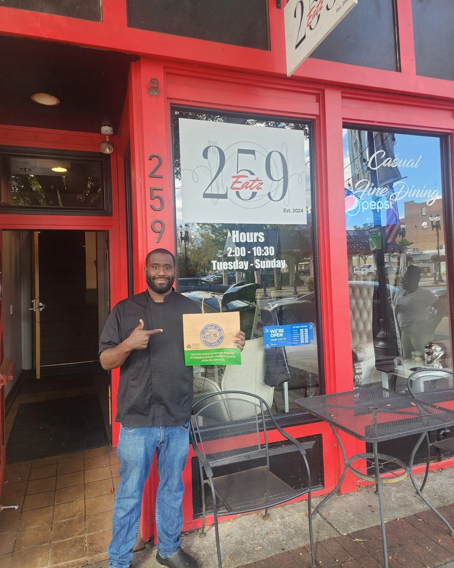 Man standing outside a red restaurant holding a paper bag pointing at it. The restaurant has large glass windows with signs displaying the name "2E9 Eatz," hours from 2:00 to 10:30, and words like 'casual' and 'fine dining.' There are chairs and smal